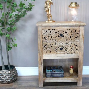 May include: A light-coloured wooden bedside table with carved floral drawer fronts. A gold statue, a glass lamp, and a stack of books sit on the table. A decorative plant is to the left.