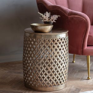 May include: A gold-coloured metal side table with a patterned design. The table has a round top and a cylindrical body with a floral cutout pattern. A small gold bowl and a vase with dried flowers sit on top. A pink armchair is in the background.
