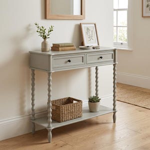 May include: A grey console table with two drawers and turned legs. A woven basket and a small potted plant sit on the lower shelf. The table is decorated with a vase of greenery, books, and a framed botanical print.