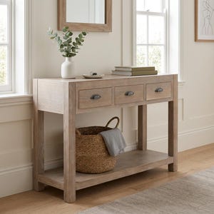 May include: A light-toned wooden console table featuring three drawers with dark metal cup pulls. The table has a lower shelf holding a woven wicker basket with a grey cloth inside. A white vase with green eucalyptus stems sits on top.