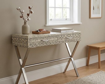 May include: A console table with a snakeskin pattern on the top and drawers, supported by a polished chrome base. The table has two drawers with decorative pulls. A vase with cotton stems, books, and a small tray with items are on top.