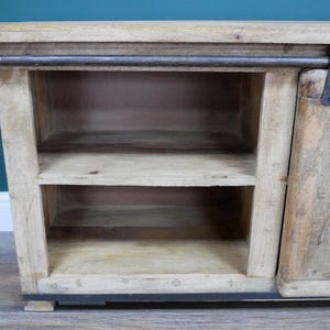 May include: A light-colored wooden cabinet with two shelves and a sliding door. The cabinet has a rustic, weathered appearance with metal hardware. The shelves are open, and the cabinet sits on a dark base.