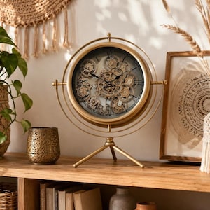 May include: A gold-toned table clock featuring an exposed gear mechanism behind a glass face. The clock is mounted within a circular frame supported by a three-legged tripod stand. It displays black Roman numerals and thin black clock hands.