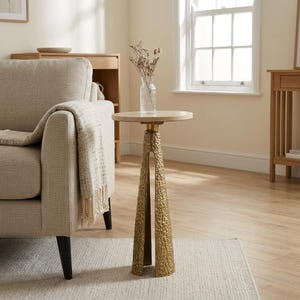 May include: A small, round side table with a textured, gold-coloured base and a light-coloured, circular top. The base has a unique, open design. The table is placed on a light-coloured rug next to a beige sofa. The table's height is approximately 61 cm.
