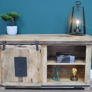 May include: A light-colored wooden cabinet with a sliding door and open shelving. A small bonsai tree in a gray pot sits on top, along with a metal lantern and a decorative lightbulb. The cabinet has a metal plate that reads "Jupiter Iron Works Est 1925."