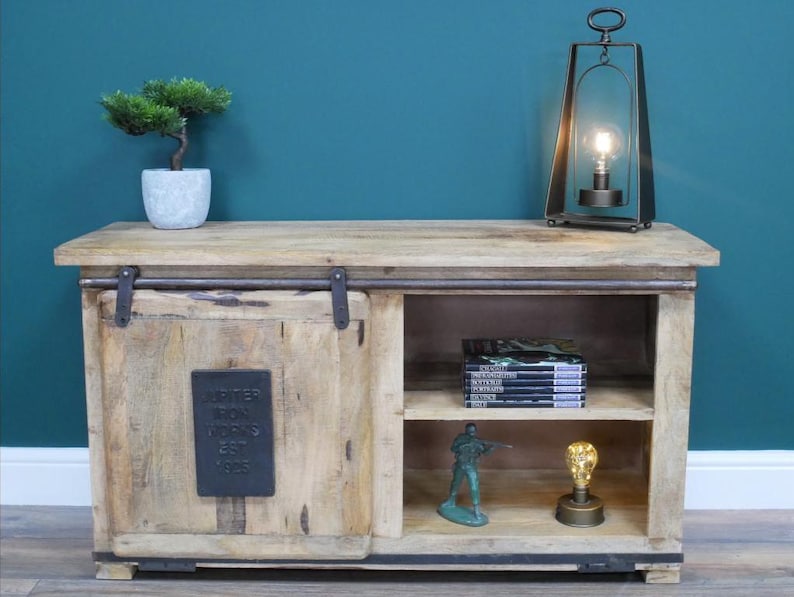 May include: A light-colored wooden cabinet with a sliding door and open shelving. A small bonsai tree in a gray pot sits on top, along with a metal lantern and a decorative lightbulb. The cabinet has a metal plate that reads "Jupiter Iron Works Est 1925."