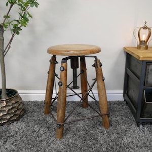 May include: A vintage-style adjustable stool featuring a circular wooden seat and four wooden legs. The legs are joined by metal supports and have metal detailing. The stool is set against a neutral backdrop, with a plant and a side table in view.