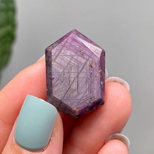 May include: A hand holds a faceted, hexagonal purple crystal. The crystal's surface is polished, revealing internal lines and inclusions. The colour varies from deep purple to lighter shades. The crystal's shape is a six-sided prism.