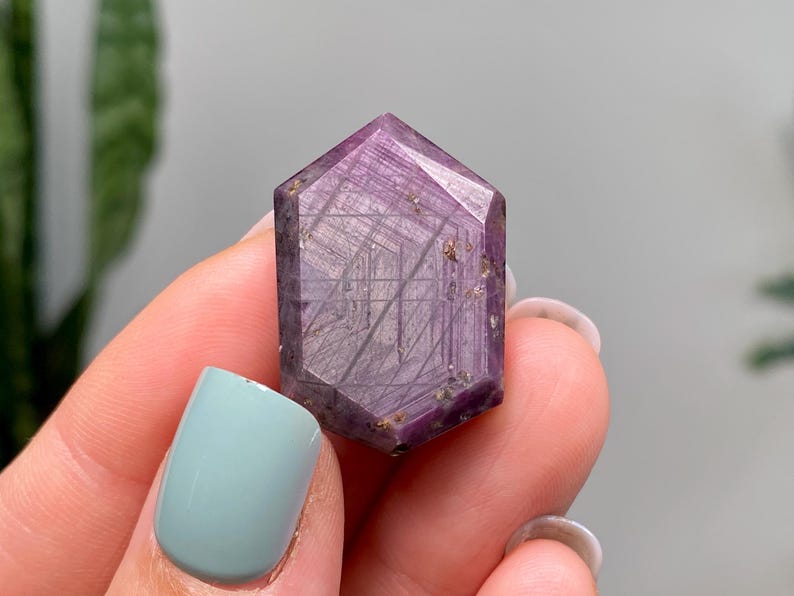 May include: A hand holds a faceted, hexagonal purple crystal. The crystal's surface is polished, revealing internal lines and inclusions. The colour varies from deep purple to lighter shades. The crystal's shape is a six-sided prism.