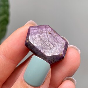 May include: A faceted, purple gemstone with a geometric shape. The stone has a network of fine lines across its surface. The gemstone is held between fingers with a light blue nail polish.