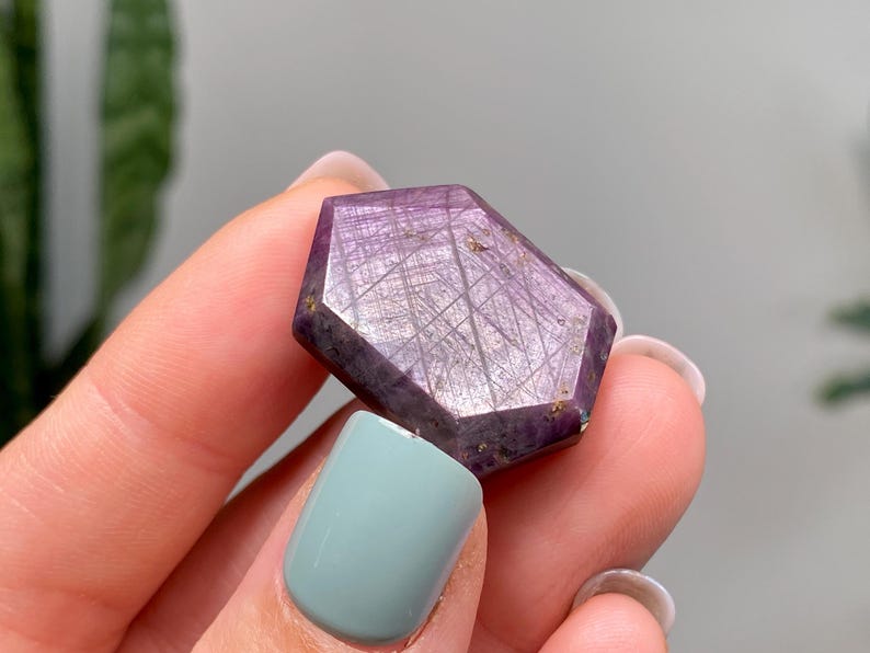 May include: A faceted, purple gemstone with a geometric shape. The stone has a network of fine lines across its surface. The gemstone is held between fingers with a light blue nail polish.
