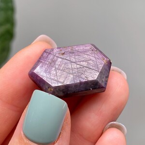 May include: A hexagonal gemstone with a deep purple colour and a shimmering, textured surface. The gem is held in a hand, showcasing its polished facets and intricate internal patterns. The stone's colour ranges from light to dark purple.