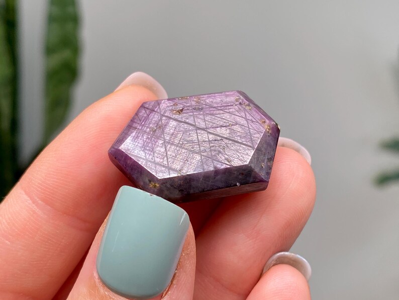 May include: A hexagonal gemstone with a deep purple colour and a shimmering, textured surface. The gem is held in a hand, showcasing its polished facets and intricate internal patterns. The stone's colour ranges from light to dark purple.