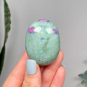 May include: Oval gemstone with a speckled, light green base and vibrant pink inclusions. The polished stone is held in a hand, showcasing its smooth texture and natural beauty. A unique mineral specimen.