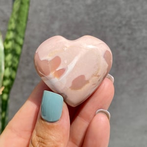 May include: A heart-shaped stone with a pink and cream colour pattern. The stone is held in a hand with a teal-coloured fingernail. The background includes a green plant and a grey wall.