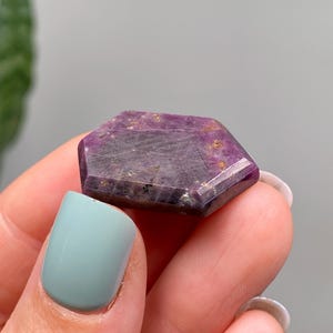 May include: A hexagonal gemstone with a deep purple colour, featuring lighter purple and gold flecks. The crystal is held in a hand with a light blue painted fingernail.