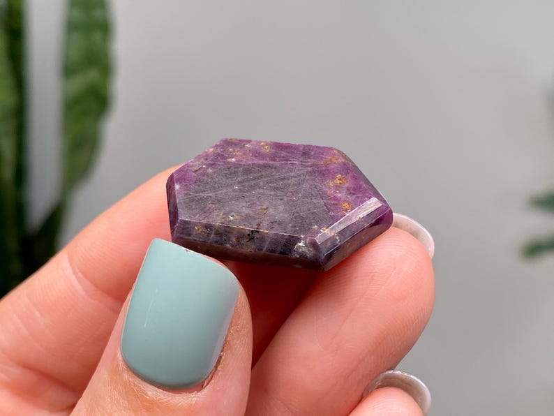 May include: A hexagonal gemstone with a deep purple colour, featuring lighter purple and gold flecks. The crystal is held in a hand with a light blue painted fingernail.