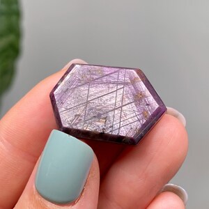 May include: A hexagonal gemstone with a deep purple colour and intricate internal patterns. The stone is held between fingers, showcasing its polished surface and unique crystalline structure. The gemstone's colour ranges from dark purple to lighter shades.