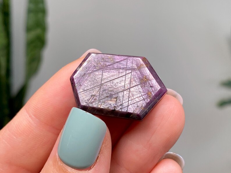 May include: A hexagonal gemstone with a deep purple colour and intricate internal patterns. The stone is held between fingers, showcasing its polished surface and unique crystalline structure. The gemstone's colour ranges from dark purple to lighter shades.