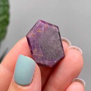 May include: A hexagonal gemstone with a faceted surface, displaying shades of purple and brown. The stone has a polished finish with visible inclusions and a subtle sheen. The gemstone is held between fingers, with light blue nail polish.