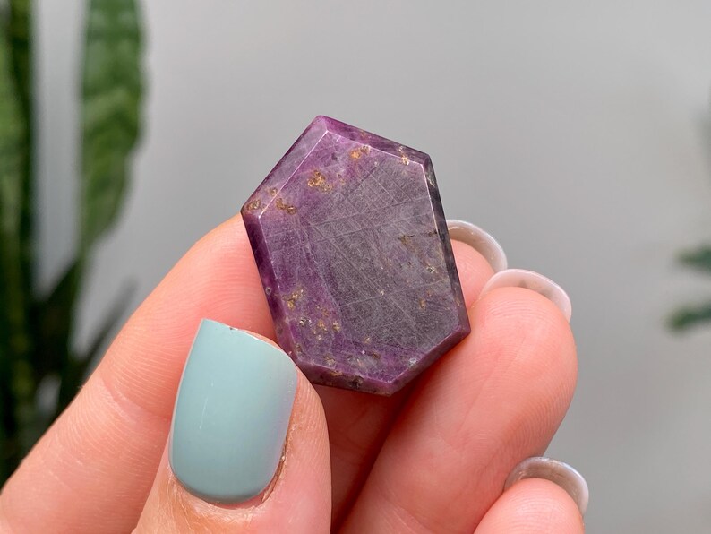 May include: A hexagonal gemstone with a faceted surface, displaying shades of purple and brown. The stone has a polished finish with visible inclusions and a subtle sheen. The gemstone is held between fingers, with light blue nail polish.