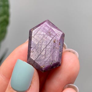 May include: A faceted, purple gemstone with a hexagonal shape. The crystal has a polished surface with visible internal striations. The gemstone is held in a hand with a light blue painted fingernail.