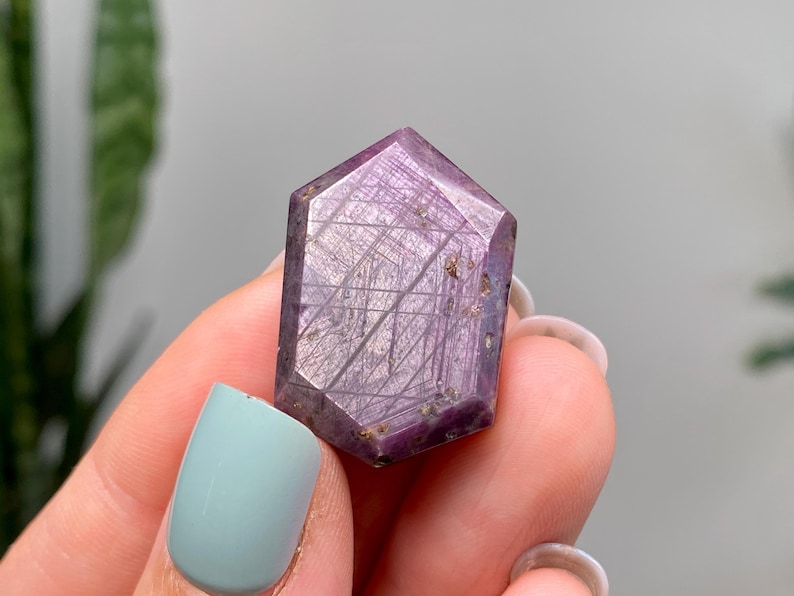 May include: A faceted, purple gemstone with a hexagonal shape. The crystal has a polished surface with visible internal striations. The gemstone is held in a hand with a light blue painted fingernail.