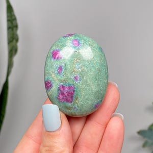 May include: An oval gemstone, polished to a smooth finish, displaying a mottled green hue with striking, irregular patches of deep pink-red. The stone is held in a hand, highlighting its reflective surface. A natural mineral specimen.