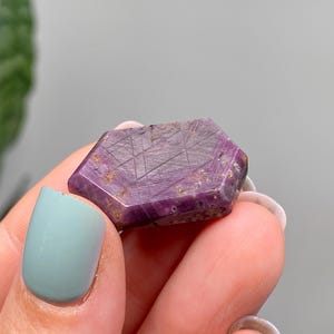 May include: A faceted, hexagonal gemstone with a deep purple hue, held between fingers. The stone has a textured surface with etched lines and flecks of gold. The gemstone is approximately 2.5 cm in length.