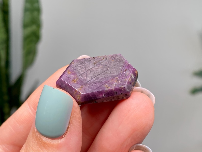 May include: A faceted, hexagonal gemstone with a deep purple hue, held between fingers. The stone has a textured surface with etched lines and flecks of gold. The gemstone is approximately 2.5 cm in length.
