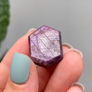 May include: A hexagonal gemstone with a deep purple colour and internal linear patterns. The stone has a polished surface. The background is blurred, with hints of green foliage. The stone appears to be held by a person.