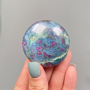 May include: A polished, circular gemstone featuring a vibrant mix of deep blue and green mineral matrix with scattered, opaque magenta-red inclusions throughout the surface, displaying a smooth, reflective finish and rounded edges.