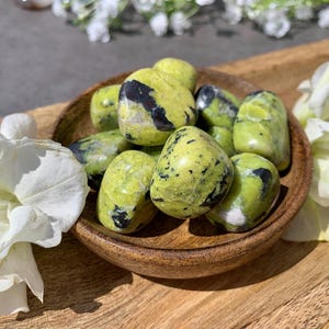May include: A collection of tumbled stones in a wooden bowl. The stones are a vibrant lime green color with black and dark green markings. White flowers and a wooden tray surround the bowl.