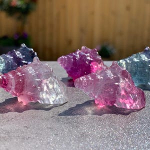 May include: Several translucent, multi-coloured crystal formations. The crystals are in shades of pink, purple, and blue. The crystals have a rough, textured surface and are arranged on a glittery surface.
