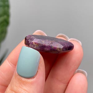 May include: A deep purple, faceted gemstone with a polished surface, held between fingers. The stone has visible inclusions. The background is blurred, with a hint of greenery and a neutral wall.