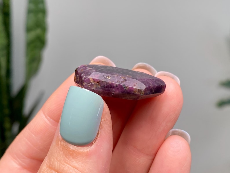 May include: A deep purple, faceted gemstone with a polished surface, held between fingers. The stone has visible inclusions. The background is blurred, with a hint of greenery and a neutral wall.