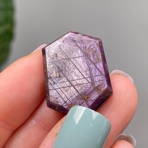 May include: A hexagonal gemstone with a deep purple colour. The stone features a network of fine, linear inclusions and small, golden-coloured inclusions. The gem is held between fingers with light blue nail polish.