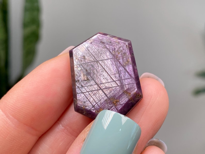 May include: A hexagonal gemstone with a deep purple colour. The stone features a network of fine, linear inclusions and small, golden-coloured inclusions. The gem is held between fingers with light blue nail polish.