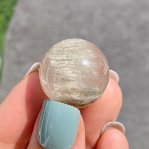 May include: A clear, spherical crystal ball with internal, light-colored inclusions. The orb is held between fingers with a light blue painted fingernail in the foreground. The background is a blurred green plant and a gray surface.