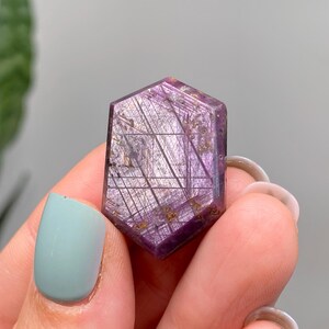 May include: A hexagonal purple gemstone with silver and gold inclusions. The gemstone is held between fingers with light blue nail polish. The gemstone has a polished surface and a geometric pattern.