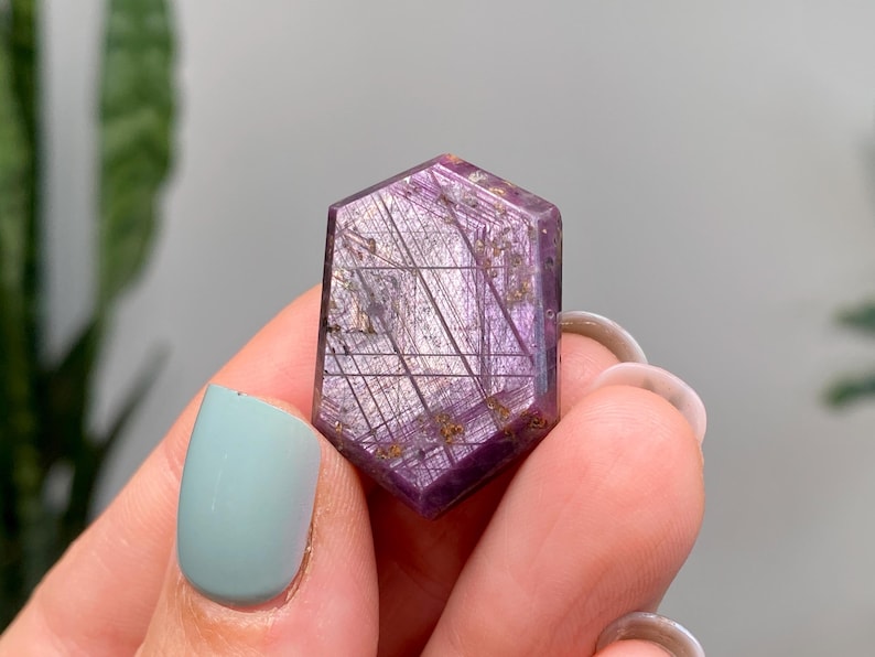 May include: A hexagonal purple gemstone with silver and gold inclusions. The gemstone is held between fingers with light blue nail polish. The gemstone has a polished surface and a geometric pattern.