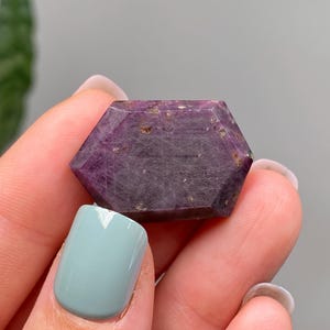 May include: A hexagonal ruby gemstone with a deep purple colour. The gem is held between fingers, showing its polished surfaces and natural inclusions. The stone's colour varies from dark purple to reddish-purple.
