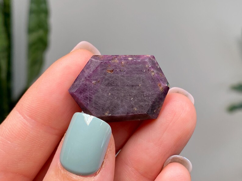 May include: A hexagonal ruby gemstone with a deep purple colour. The gem is held between fingers, showing its polished surfaces and natural inclusions. The stone's colour varies from dark purple to reddish-purple.