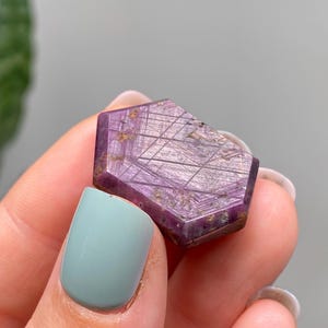 May include: A faceted, purple gemstone with a geometric pattern and gold flecks. The gemstone is held between fingers with a light blue painted fingernail.