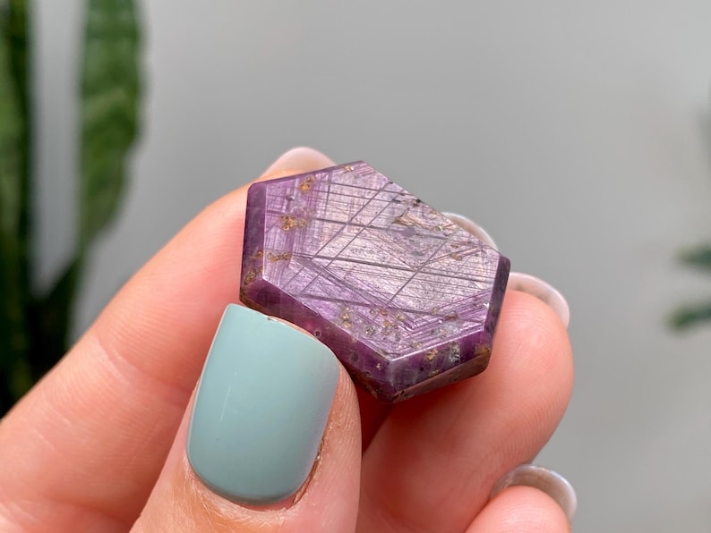 May include: A faceted, purple gemstone with a geometric pattern and gold flecks. The gemstone is held between fingers with a light blue painted fingernail.