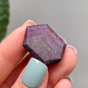 May include: A faceted, purple gemstone with a geometric shape. The stone has a deep, rich colour with visible internal patterns and inclusions. The gemstone is held between fingers with a light blue nail polish.