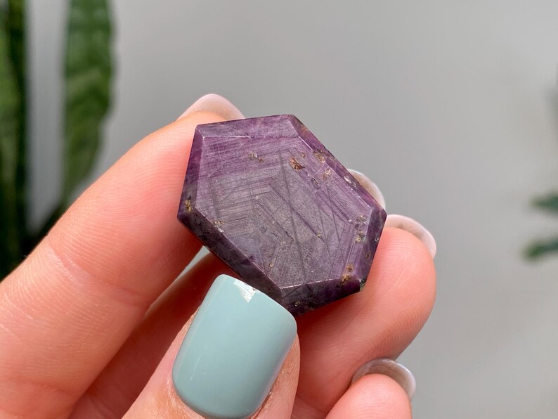May include: A faceted, purple gemstone with a geometric shape. The stone has a deep, rich colour with visible internal patterns and inclusions. The gemstone is held between fingers with a light blue nail polish.