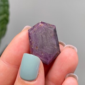May include: A hexagonal crystal with a deep purple colour and internal inclusions. The crystal is held between fingers, showing its polished surfaces and natural beauty. The gemstone's colour ranges from light to dark purple.