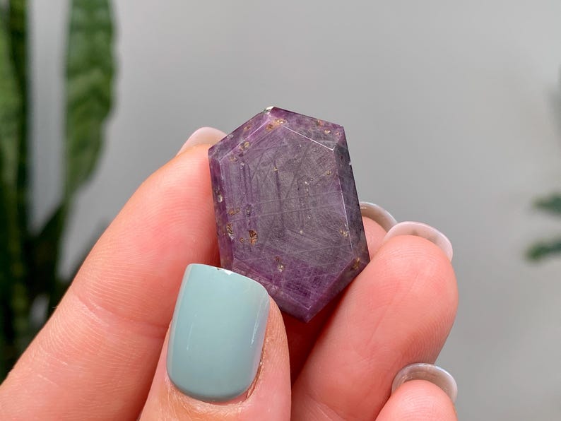 May include: A hexagonal crystal with a deep purple colour and internal inclusions. The crystal is held between fingers, showing its polished surfaces and natural beauty. The gemstone's colour ranges from light to dark purple.