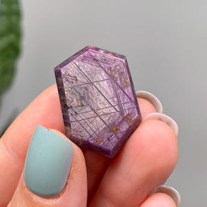 May include: A hexagonal gemstone with a deep purple colour. The stone has intricate, linear inclusions in gold and silver tones, creating a unique pattern. The gemstone is held between fingers.
